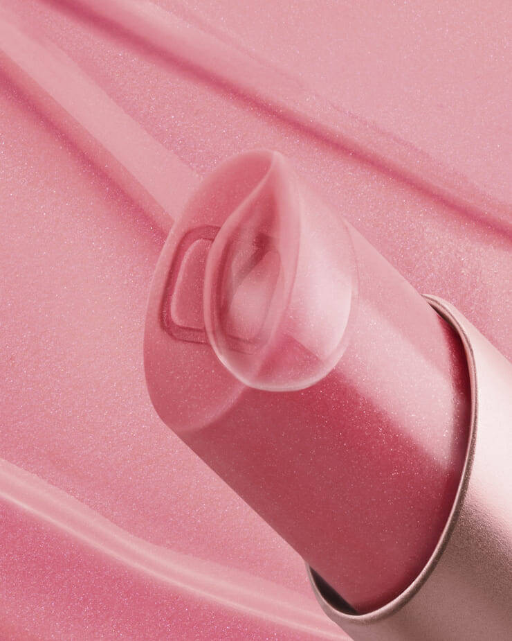 A close-up shot of a pink lip balm, with its clear, translucent cap angled slightly open, revealing a subtle "V" logo inside, all set against a soft pink background with faint streaks of product.