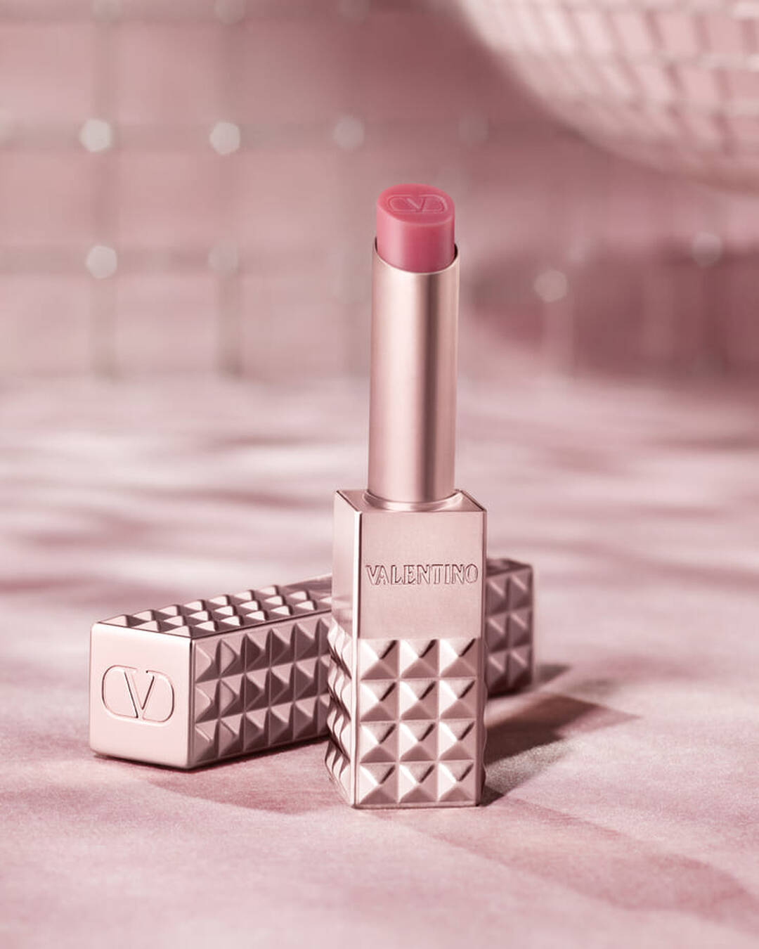 A rose gold-toned Valentino Spike lip product with a pink tip stands upright, with its matching studded case lying next to it. Both items are positioned on a soft, glittering pink background that evokes a sense of glamour.