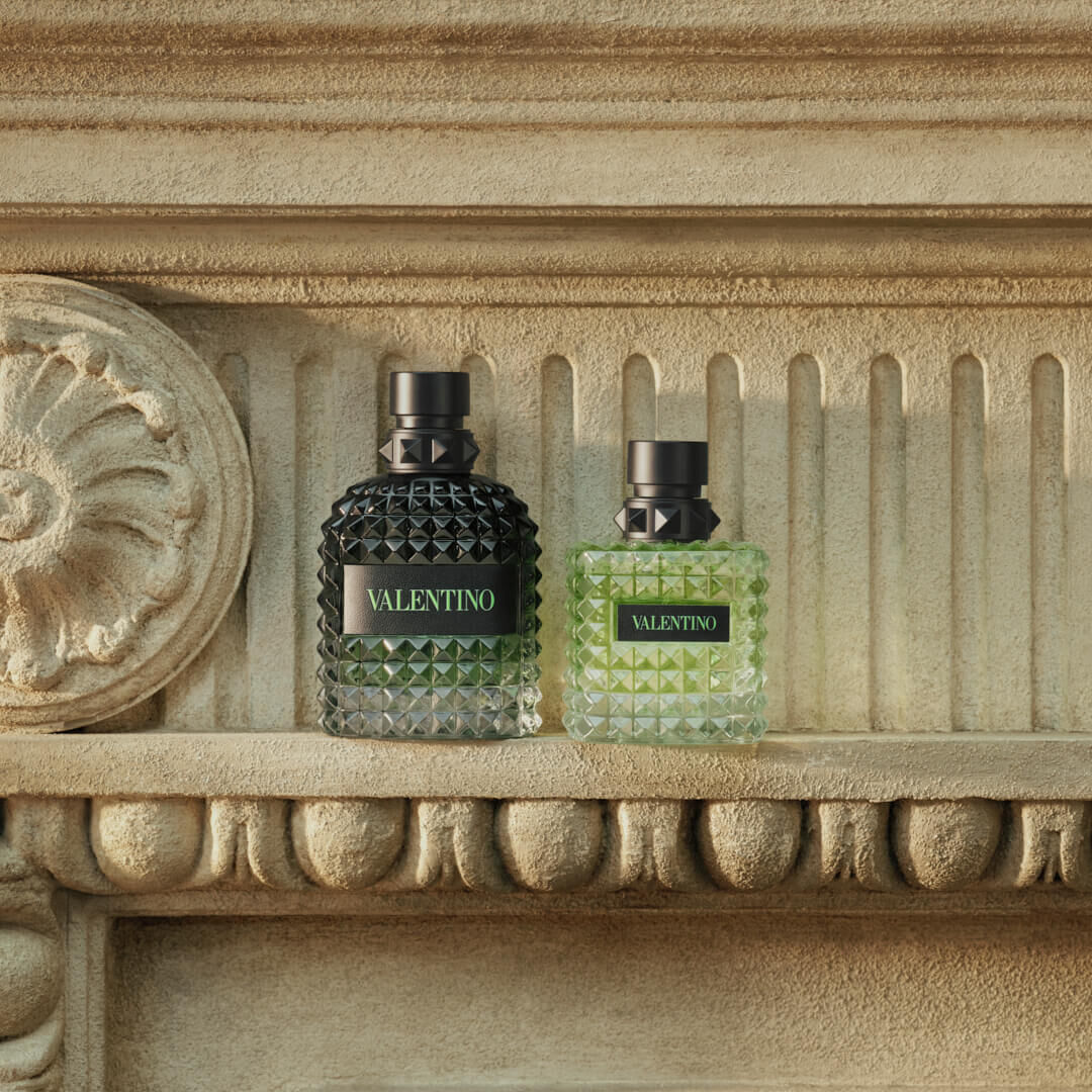 A model in a vibrant green shirt holds a Valentino perfume bottle, posing against a historic outdoor backdrop.
