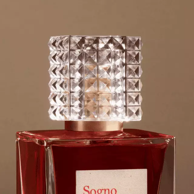 A close-up of a perfume bottle with a faceted crystal cap and a rectangular glass body filled with dark red liquid. The label reads "Sogno."