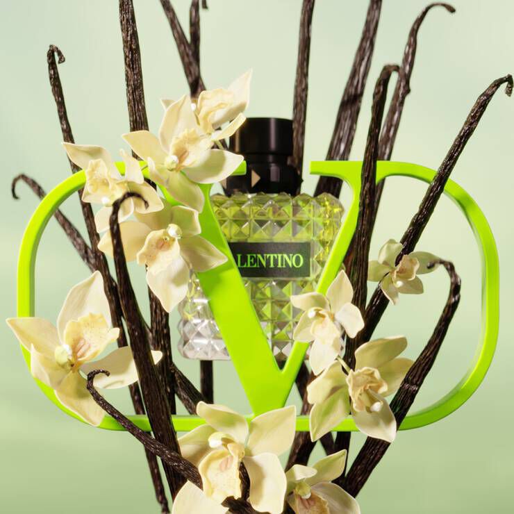 A bottle of Valentino perfume is surrounded by elegant white orchids and dark, twisted branches, set against a soft green background. The bottle is framed by a bold green logo resembling the letter "V."