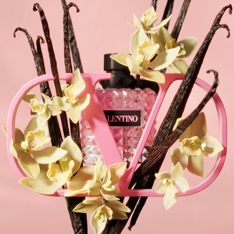 A pink Valentino perfume bottle is elegantly displayed within a stylish pink frame, surrounded by vanilla pods and delicate yellow flowers against a soft pink background.