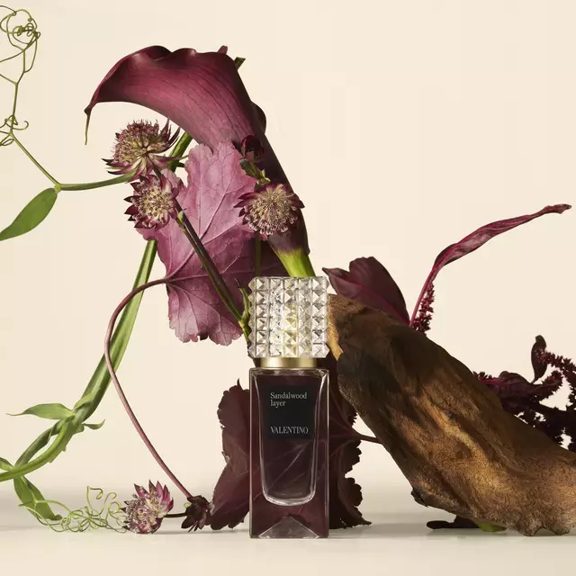 A Valentino perfume bottle placed beside deep red flowers and textured natural elements against a soft, light background.