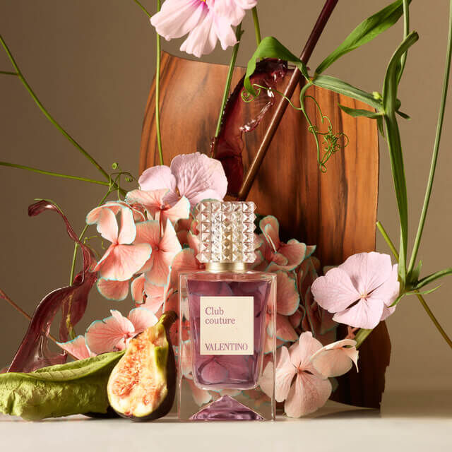 A bottle of Valentino's "Club Couture" perfume surrounded by vibrant pink flowers, a fig, and greenery on a soft background.