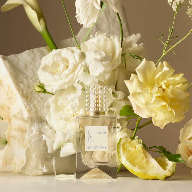 A bottle of Valentino perfume titled "L'Innocence de Fair" is positioned among light-colored flowers and a lemon slice, creating an elegant and fresh aesthetic.
