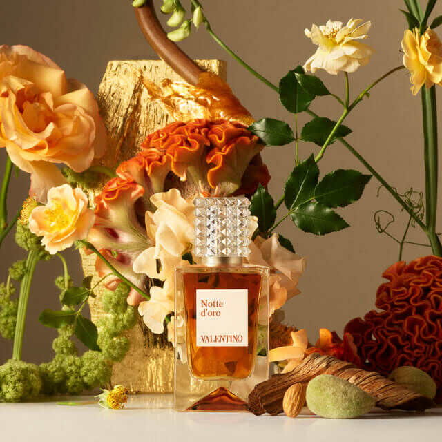 A bottle of Valentino's Notte d'Oro perfume, surrounded by vibrant flowers, textured orange fabric, and decorative elements, set against a neutral background.