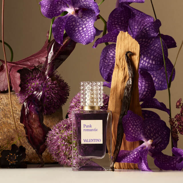 A bottle of Valentino's "Punk Romantic" perfume surrounded by purple orchids, textured leaves, and a piece of wood, creating a vibrant floral composition.