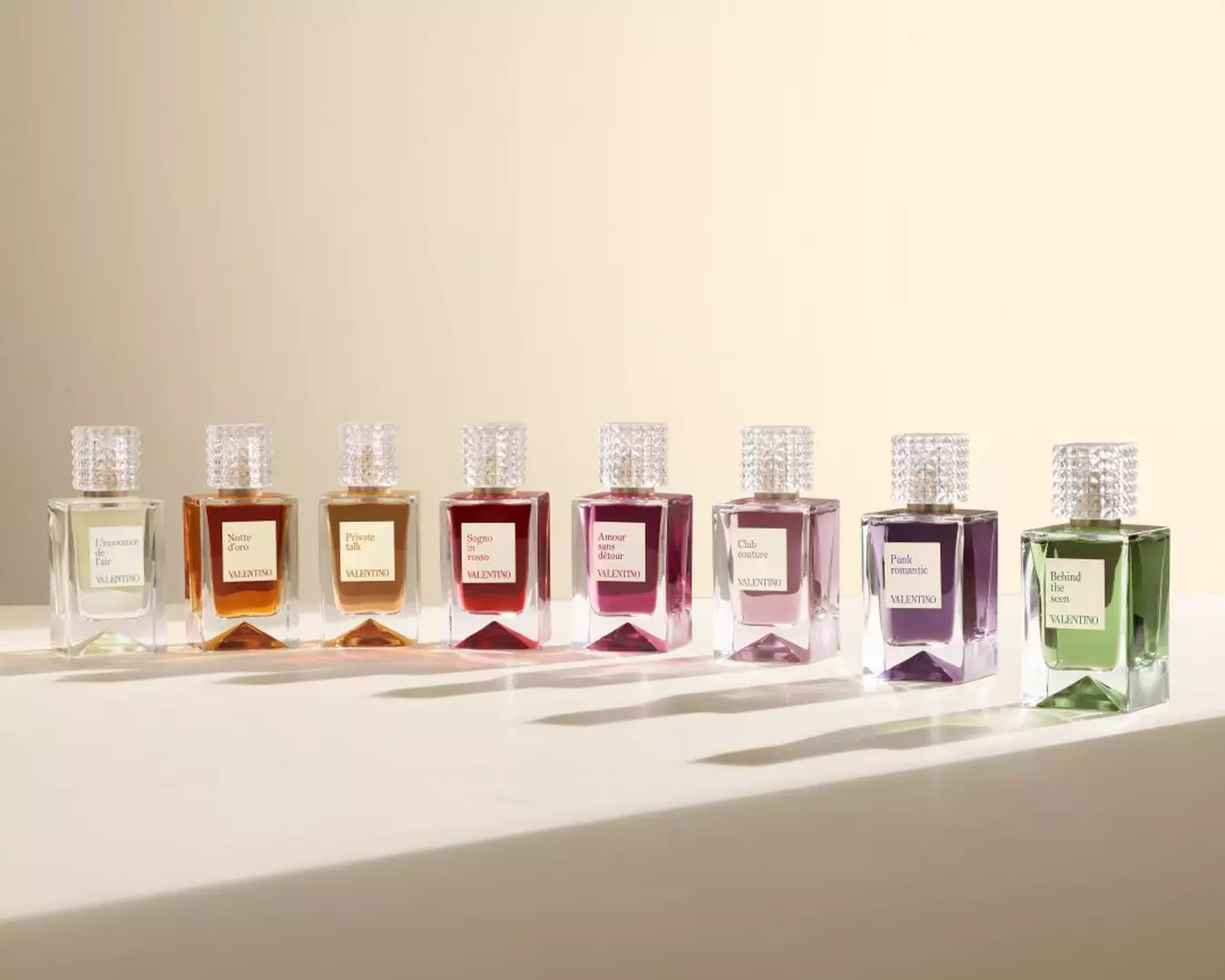 A row of eight Valentino perfume bottles in different colors, each with a textured transparent cap, arranged on a light surface against a beige background.