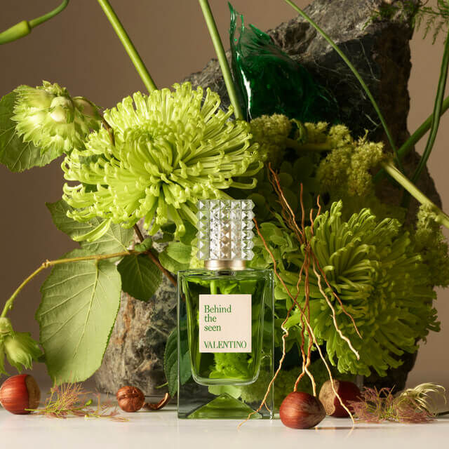 A green perfume bottle labeled "Behind the Seen" by Valentino is surrounded by various green flowers and natural elements, set against a textured stone backdrop.
