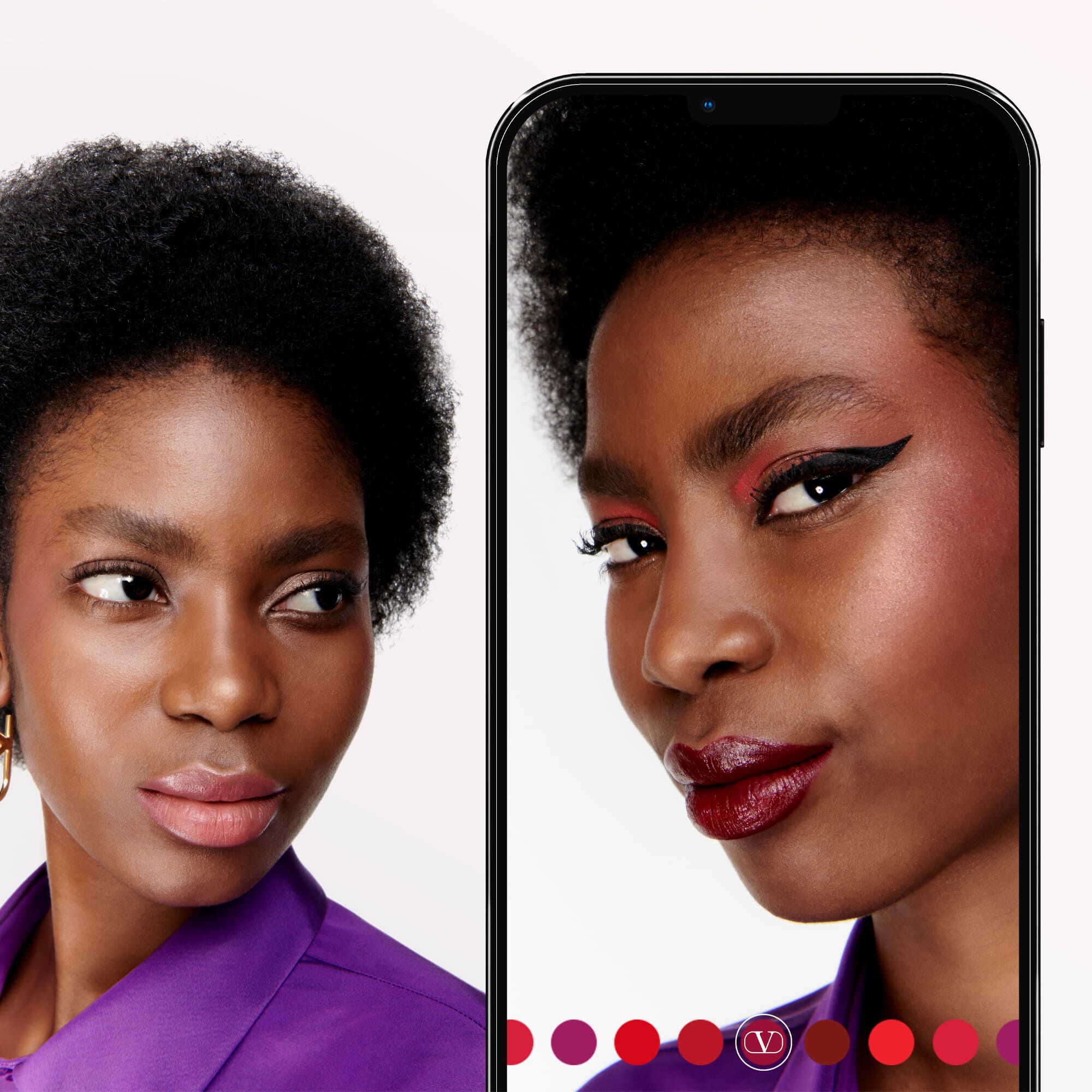 A close-up of a woman with natural textured hair, showcasing bold makeup on her eyes and lips. She is looking towards the side, with her reflection displayed on a smartphone beside her.
