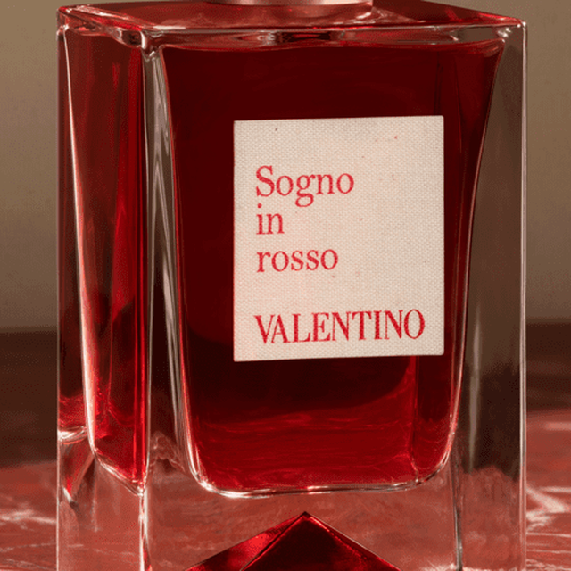 A close-up of a red glass perfume bottle with a square design and a label that reads "Sogno in rosso VALENTINO."
