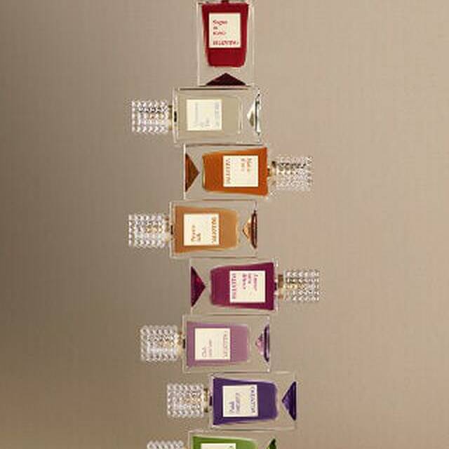 A row of eight Valentino perfume bottles in different colors, each with a textured transparent cap, arranged on a light surface against a beige background.