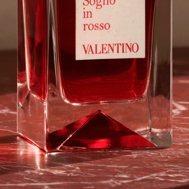 A close-up of a red perfume bottle with a clear glass base, featuring the label "Sogno in rosso" and "VALENTINO." The background has a textured, reddish surface.