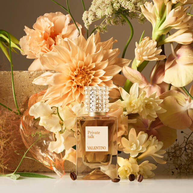 A bottle of Valentino's "Private Talk" perfume is elegantly positioned among soft, peach-colored flowers and delicate greenery, creating a fresh and sophisticated floral arrangement.