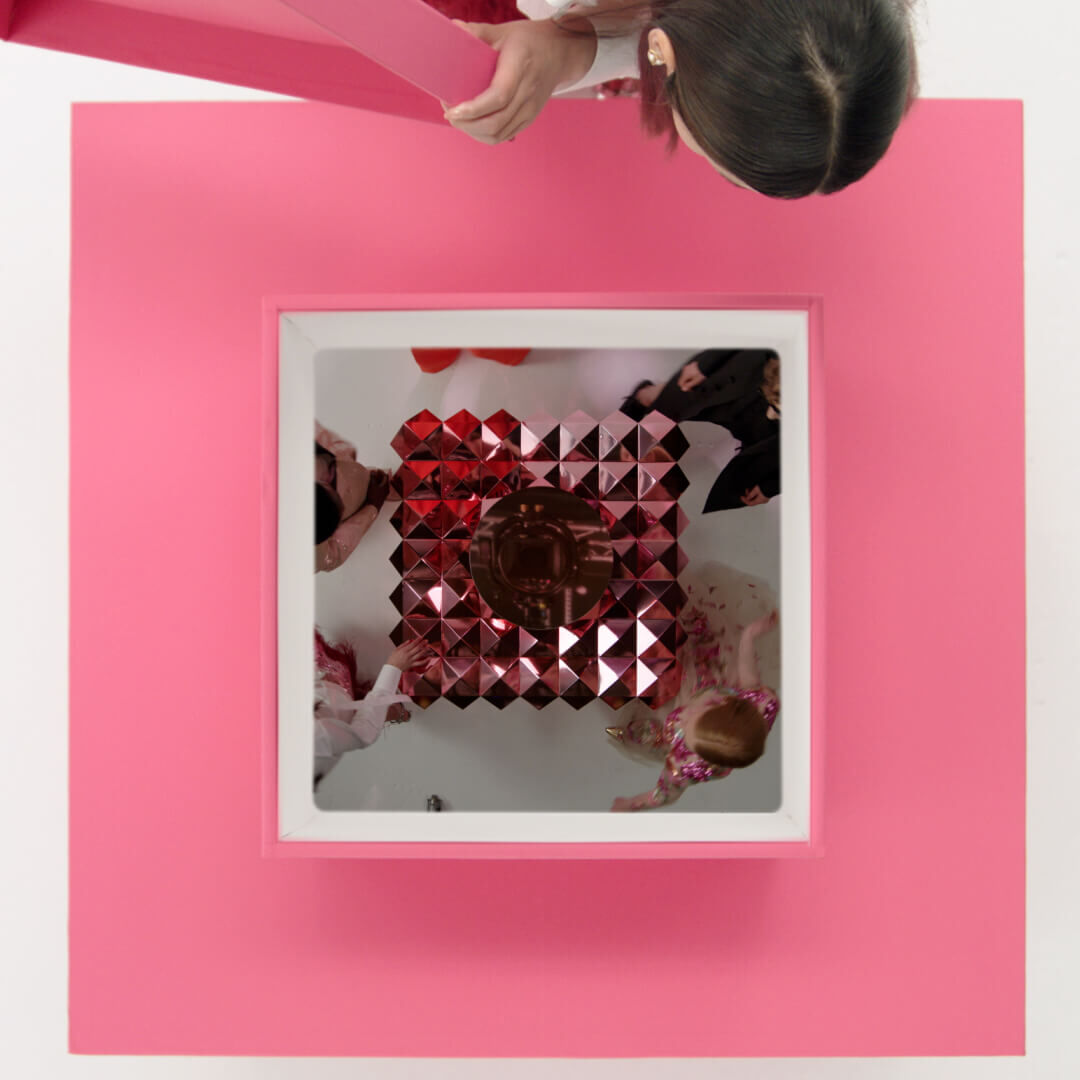 A top view of a person draping a pink fabric over a square structure, which contains a reflective surface and red geometric shapes inside. The surrounding area is minimalistic with a white background.