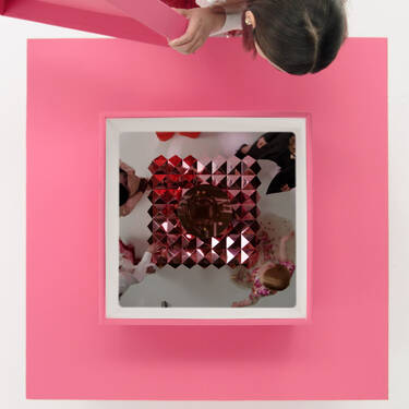A top view of a person draping a pink fabric over a square structure, which contains a reflective surface and red geometric shapes inside. The surrounding area is minimalistic with a white background.