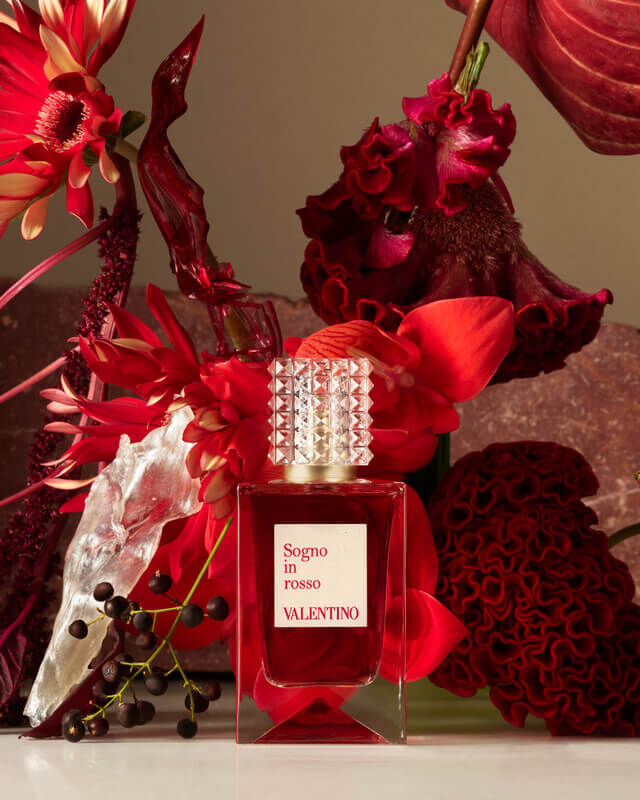 A vibrant display featuring a bottle of Valentino's Signore fragrance surrounded by an assortment of red flowers, including roses and other foliage, creating a rich and luxurious composition.